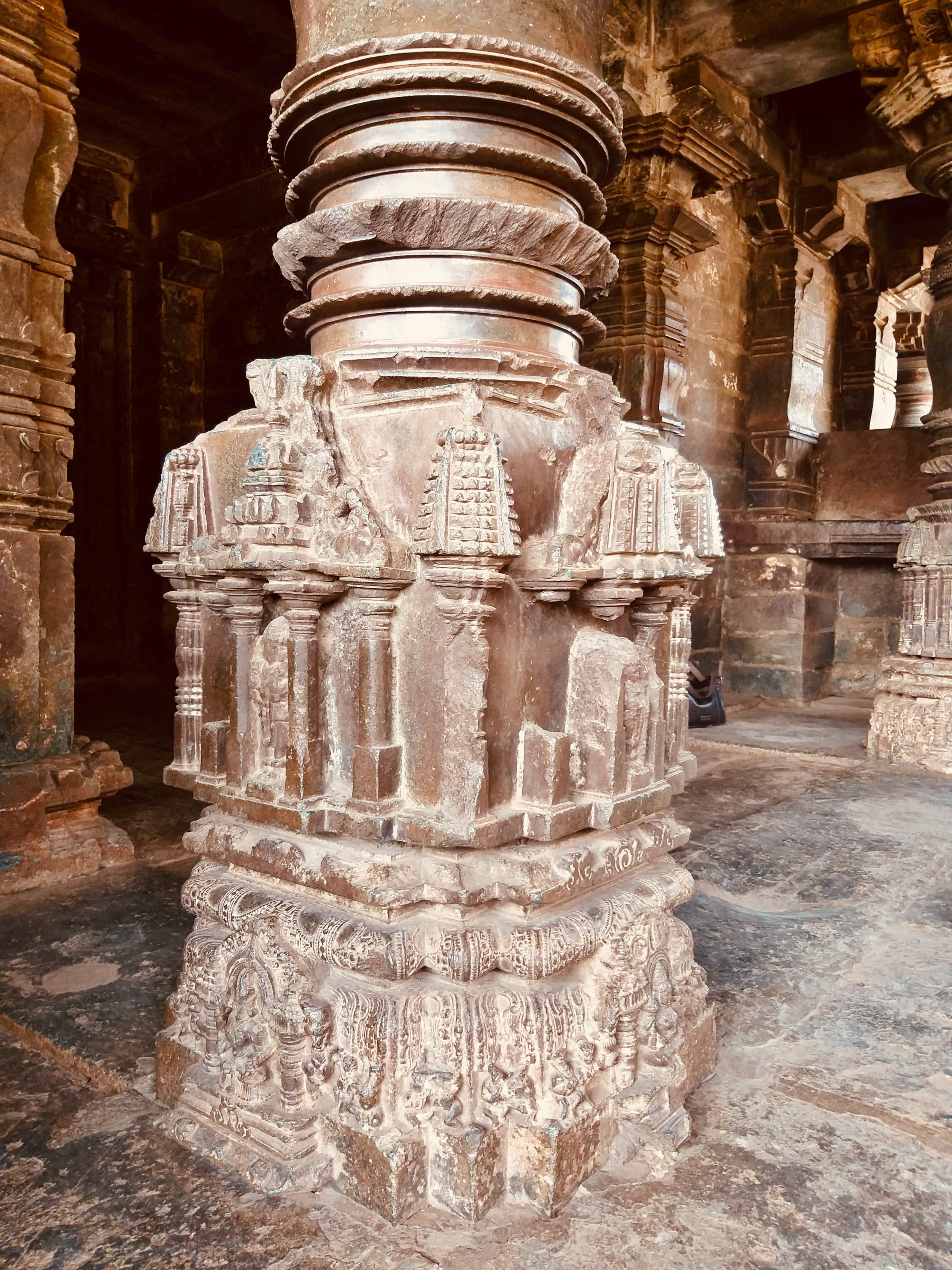 A richly carved stone pillar in a dim pillared hall, its base cut into tiers of miniature architectural detail.