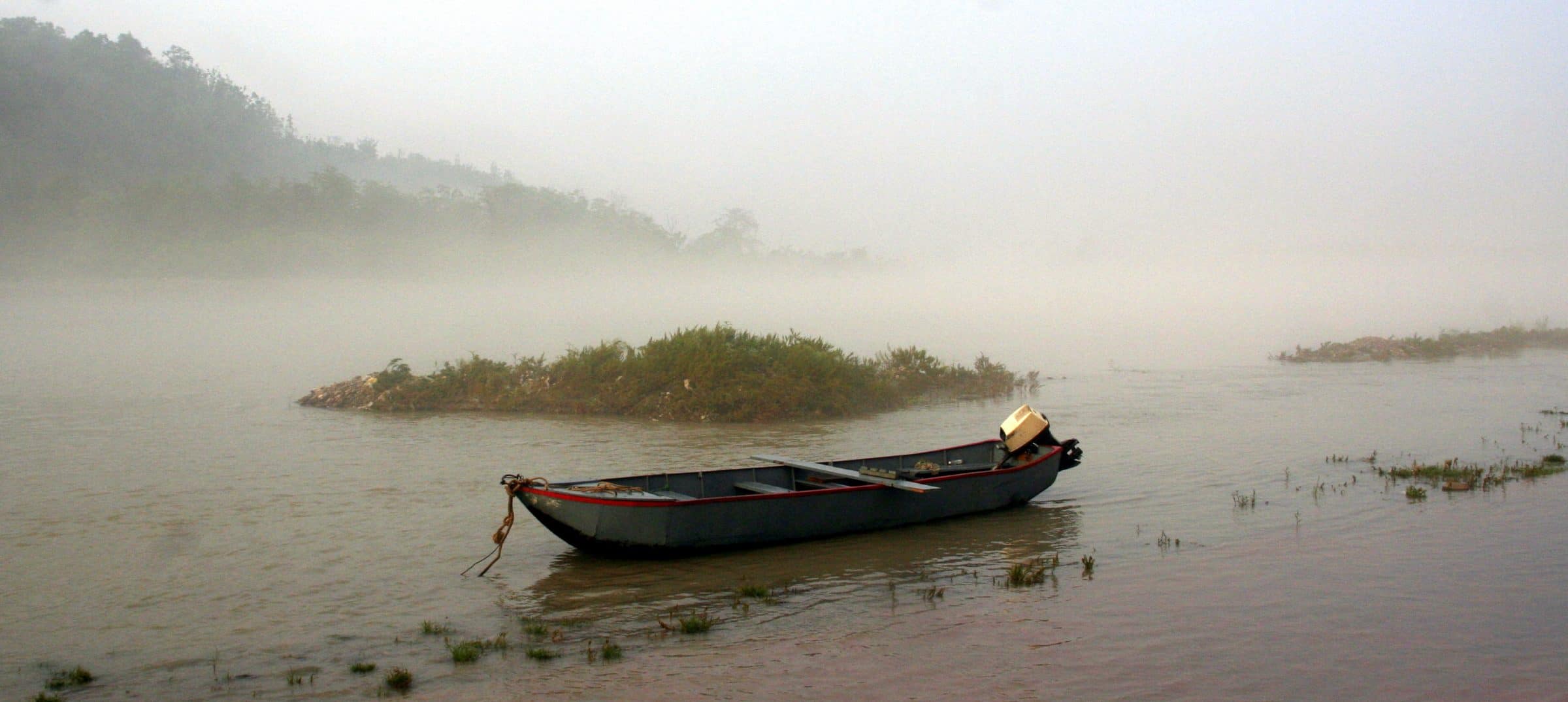A wooden boat moored on still water in heavy morning mist, with a low bank behind it.